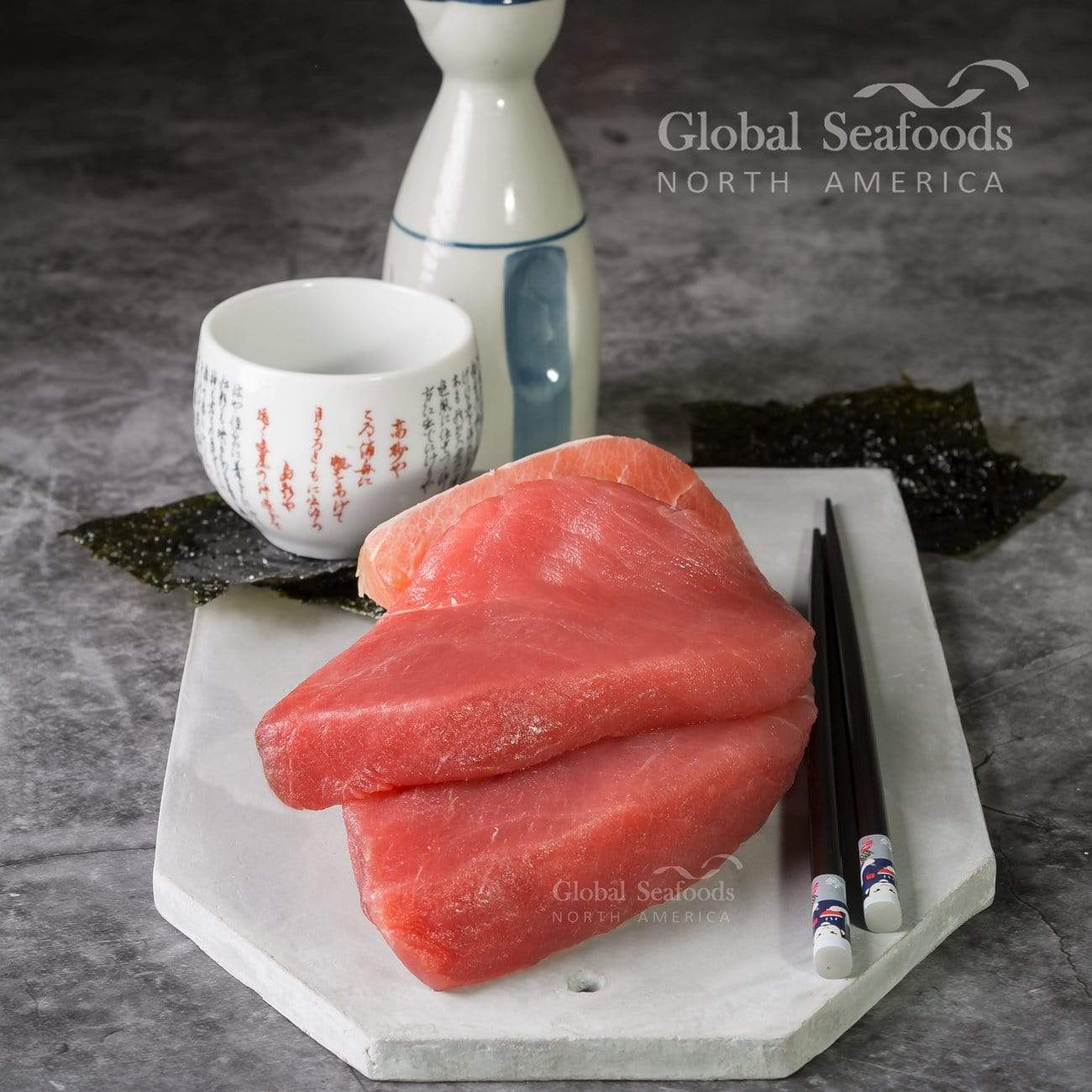 Two yellowfin tuna steaks placed on white stone, next to a sake bottle and cup, with chopsticks on the side for a refined Japanese-style presentation