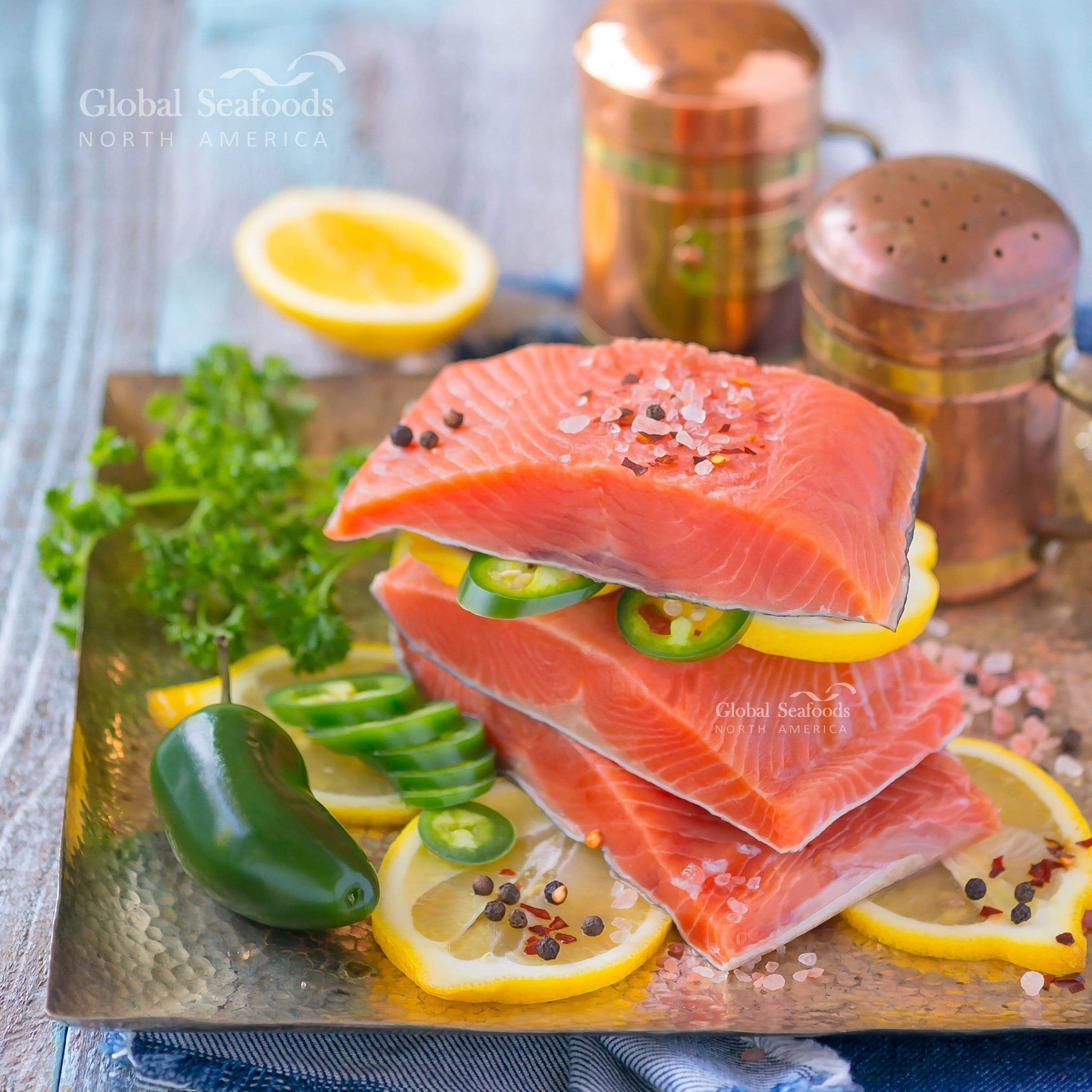 Three portions of king salmon fillets with skin on, stacked with one on top of the others, garnished with lemon slices and green chili, and topped with salt and pepper for a bold and fresh seafood presentation