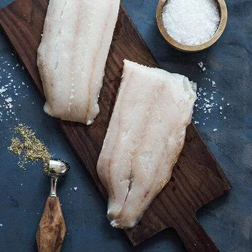 Two Lingcod fillets placed on a wooden board with a small cup of white salt beside them, styled for a clean and rustic seafood preparation